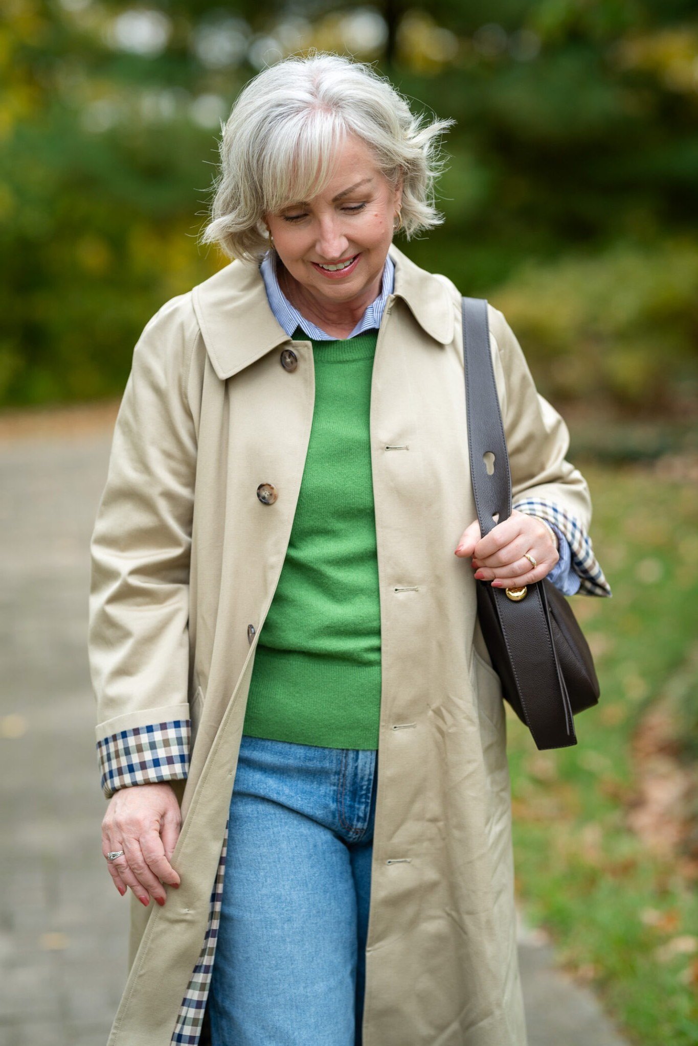 Beige trench coat, green knit sweater, blue jeans, woman on leafy path