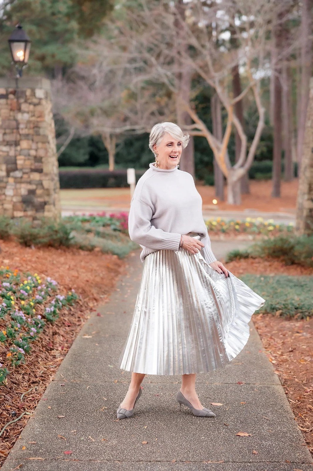 Light gray sweater, silver pleated midi skirt, woman twirling on garden path
