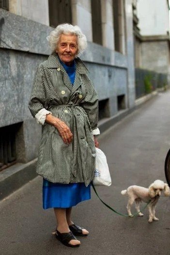 Older woman in gray trench coat over blue dress walking a dog