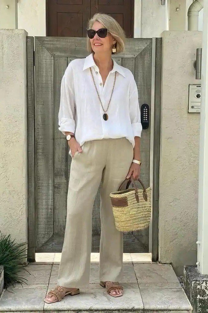 Older woman in white linen shirt and beige wide pants with straw bag