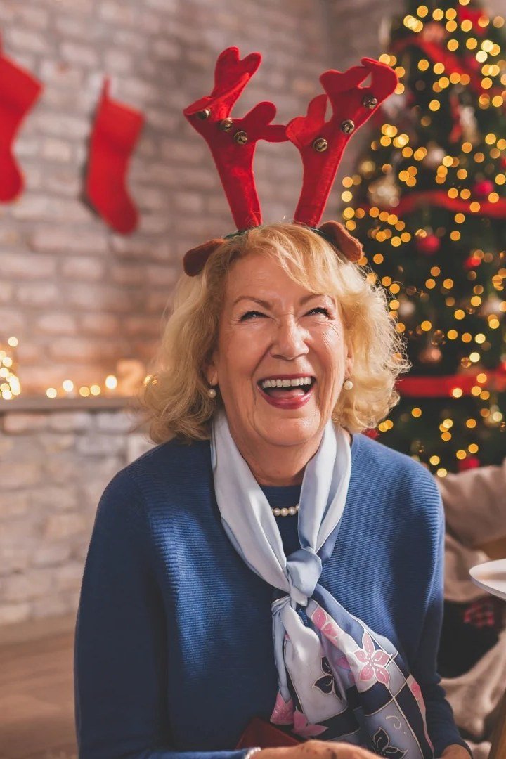 Blonde Curls With Reindeer Antlers by Christmas Tree - Christmas party hairstyle for women over 60 (blonde curls with reindeer antlers by christmas tree, front view)
