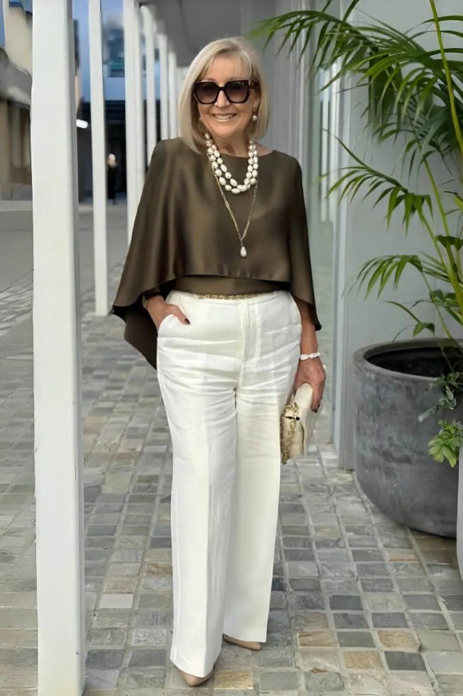 Woman over 50 wearing an olive cape-sleeve top with white wide-leg trousers