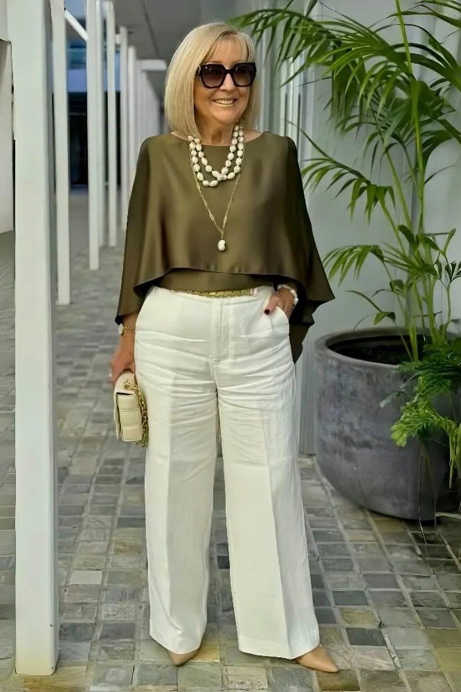 Woman over 50 wearing an olive draped top with white trousers and pearls