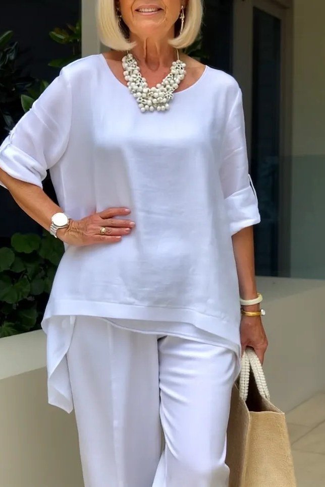 Woman over 50 wearing a white tunic with white trousers and bold pearl necklace