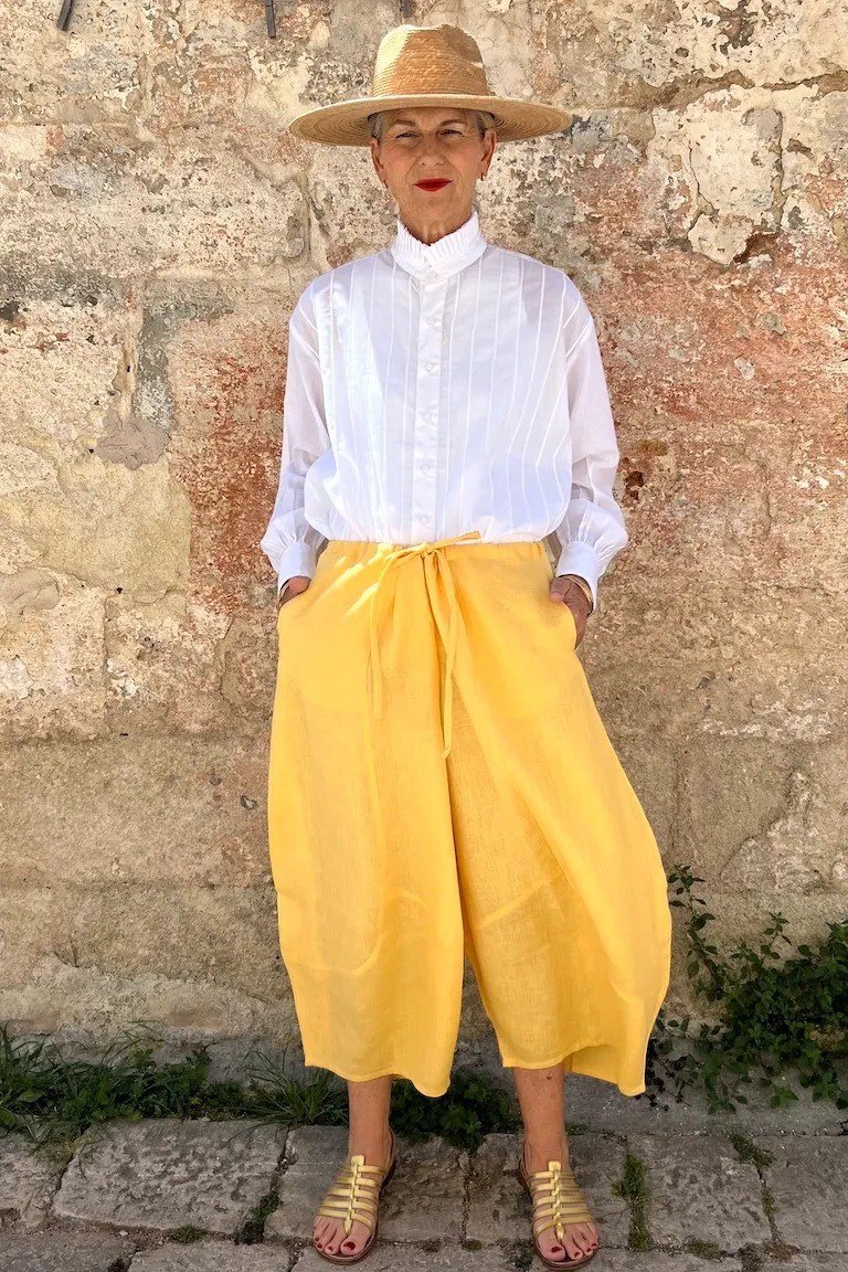 Woman over 50 in a white shirt with bright yellow wide culottes and straw hat
