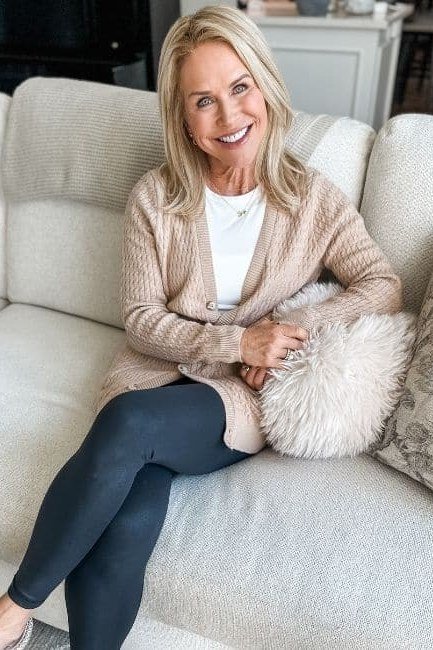 Beige knit cardigan over white top, dark leggings, cream couch, leggings outfit idea for women over 50