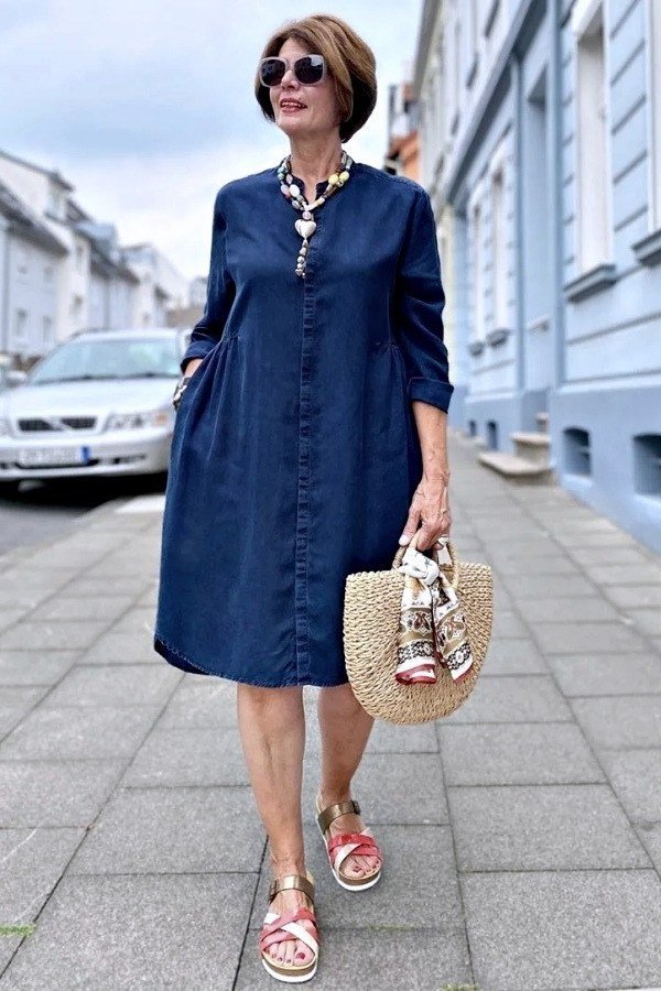 linen summer dresses for women over 60: older woman in denim-blue shirt dress with straw tote and pink sandals by colorful houses