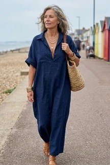 linen summer dresses for women over 60: older woman in navy v-neck maxi shirt dress with woven straw bag on beach boardwalk