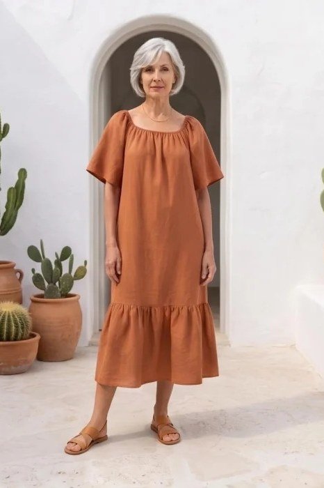 linen summer dresses for women over 60: older woman in rust off-shoulder ruffle midi dress on older white-haired woman by white wall with cacti