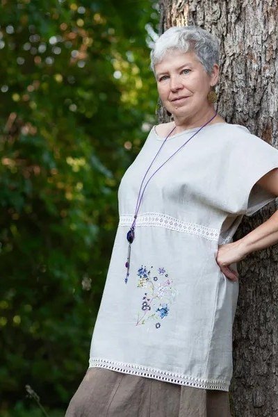 linen summer dresses for women over 60: older woman in white embroidered linen tunic midi dress with lavender necklace by tree