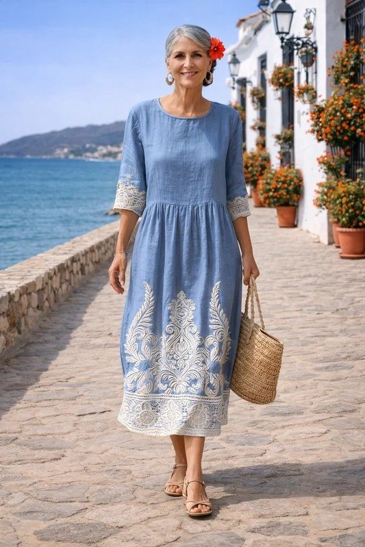 linen summer dresses for women over 60: older woman in light blue embroidered linen midi dress with straw tote by the sea