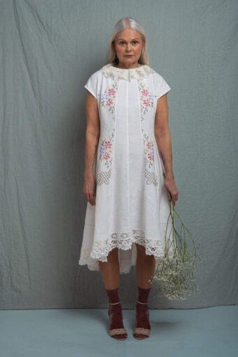 linen summer dresses for women over 60: older woman in white embroidered linen shift dress with brown boots in studio