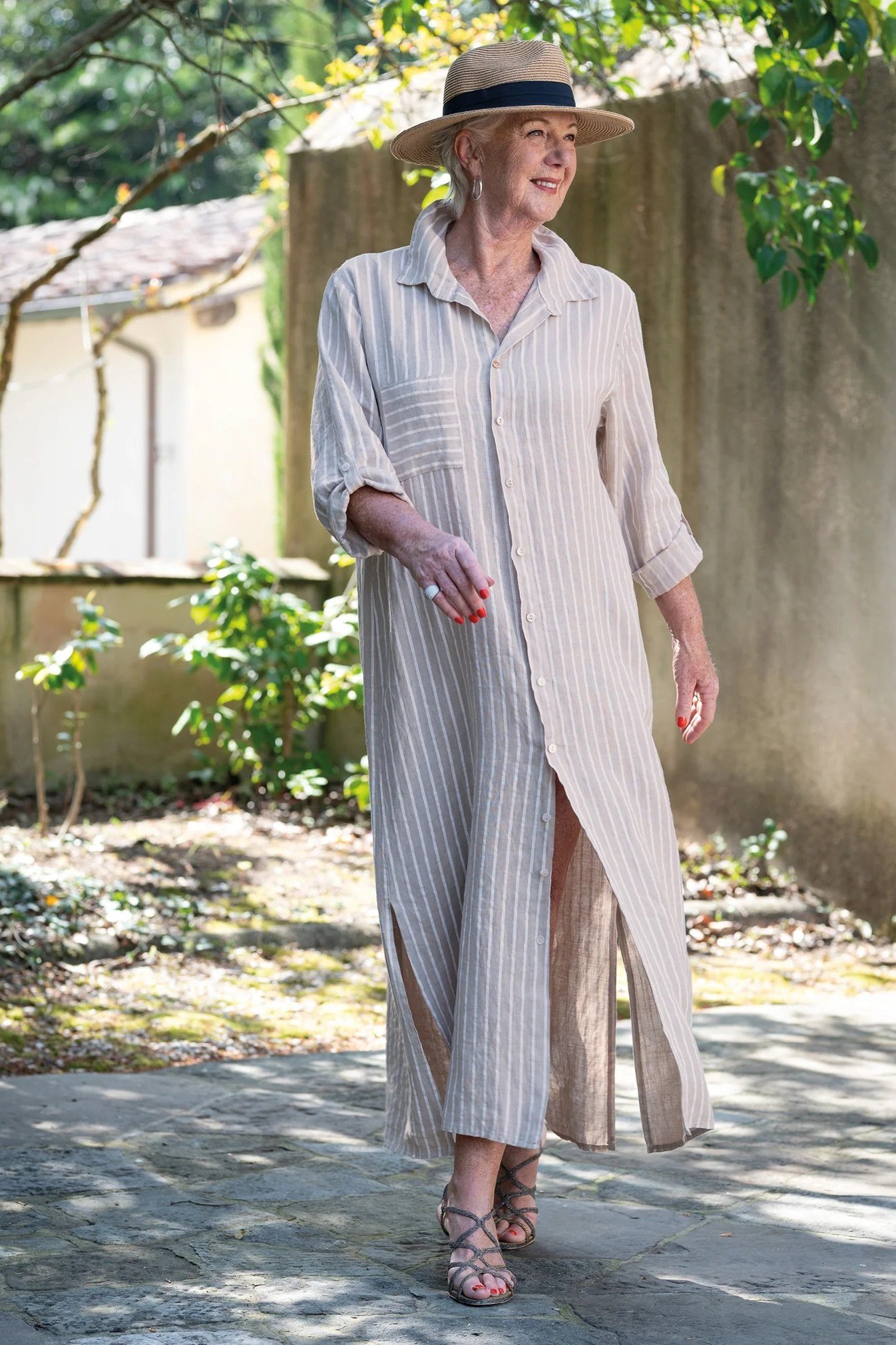 linen summer dresses for women over 60: older woman in white pinstripe linen shirt dress maxi with slit and straw hat in garden