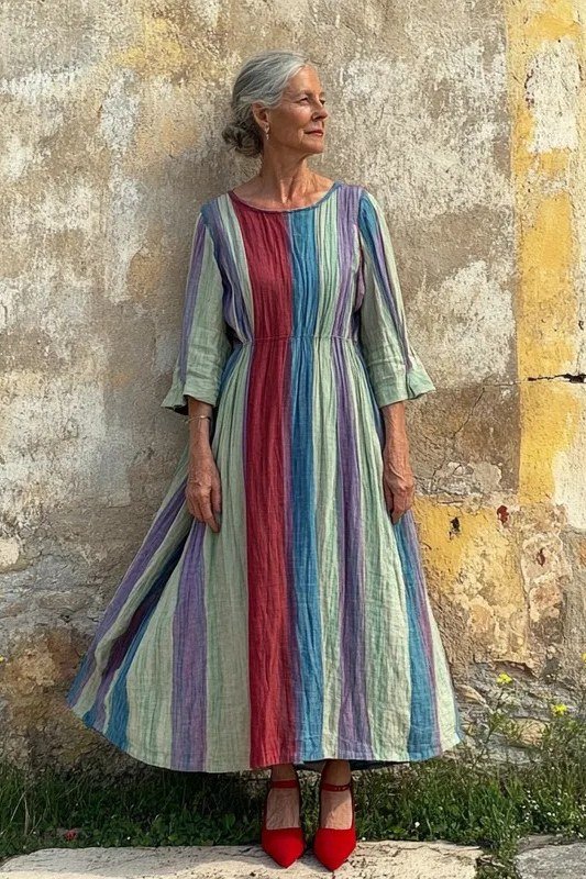 linen summer dresses for women over 60: older woman in multicolor striped three-quarter-sleeve linen midi dress with red heels by rustic wall