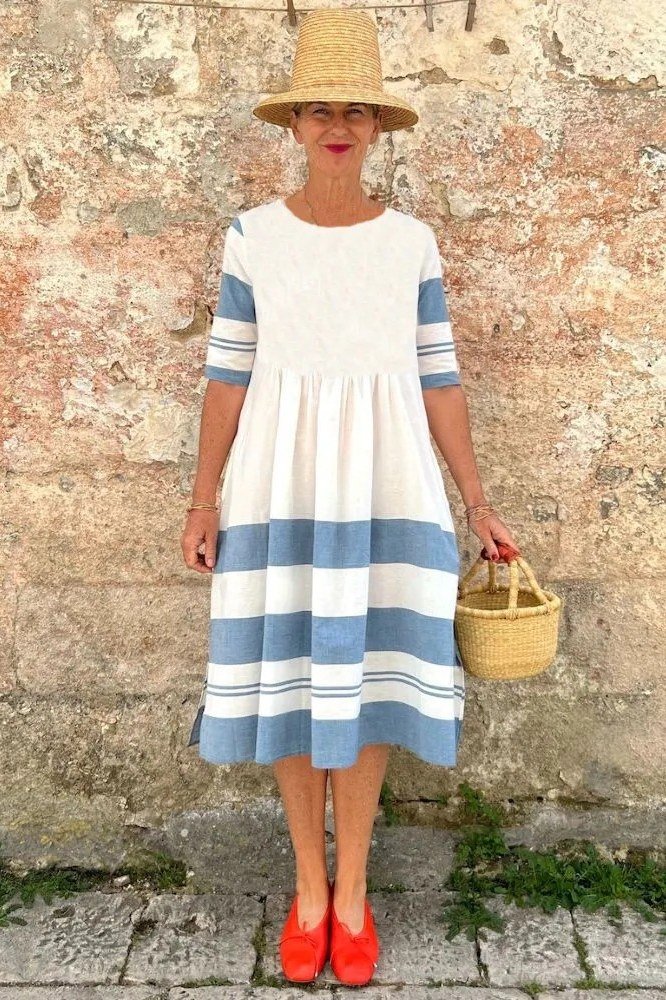 linen summer dresses for women over 60: older woman in blue and white striped linen midi dress with red heels and straw bag by stone wall