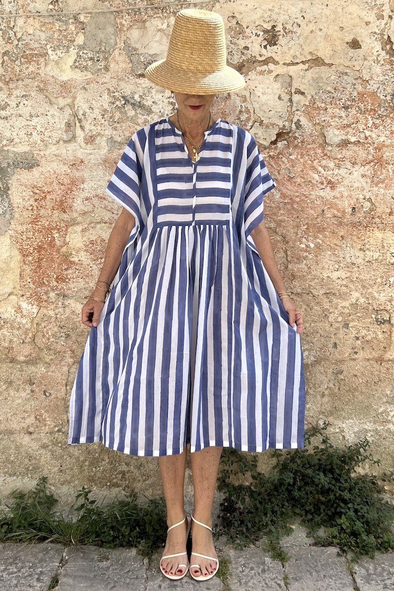 linen summer dresses for women over 60: older woman in blue and white striped short-sleeve linen midi dress with straw hat by stone wall