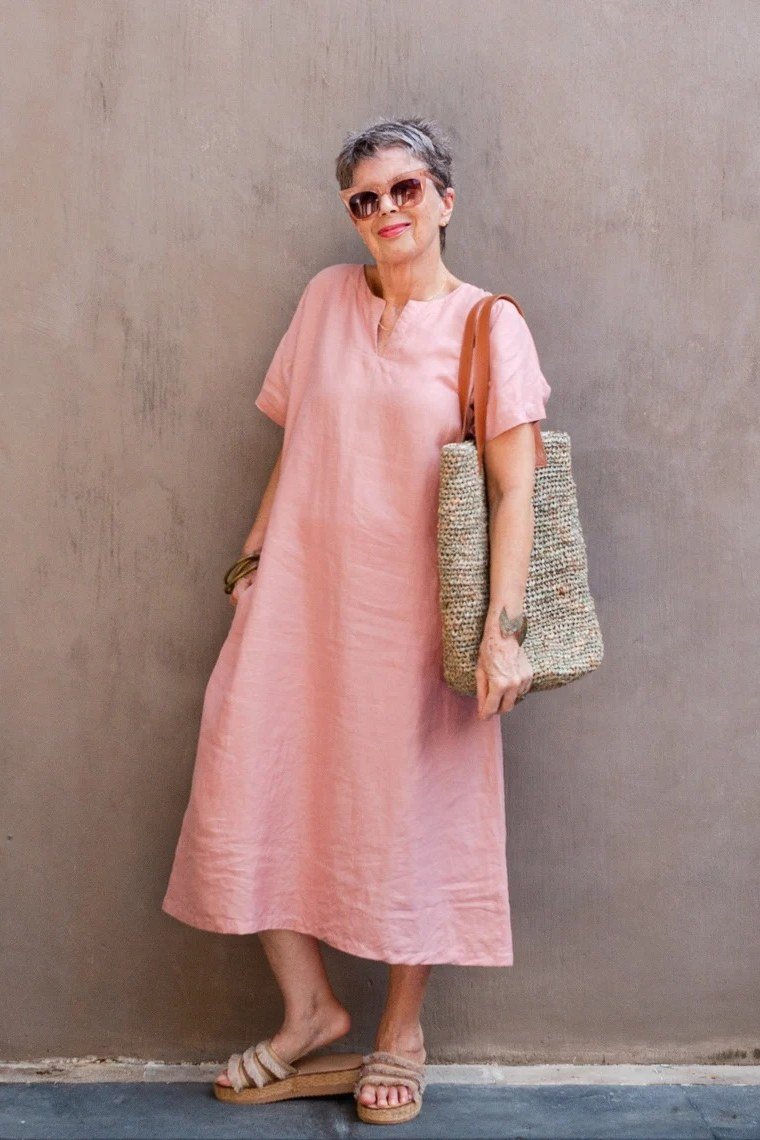 linen summer dresses for women over 60: older woman in pink short-sleeve linen midi dress with straw tote by pink wall