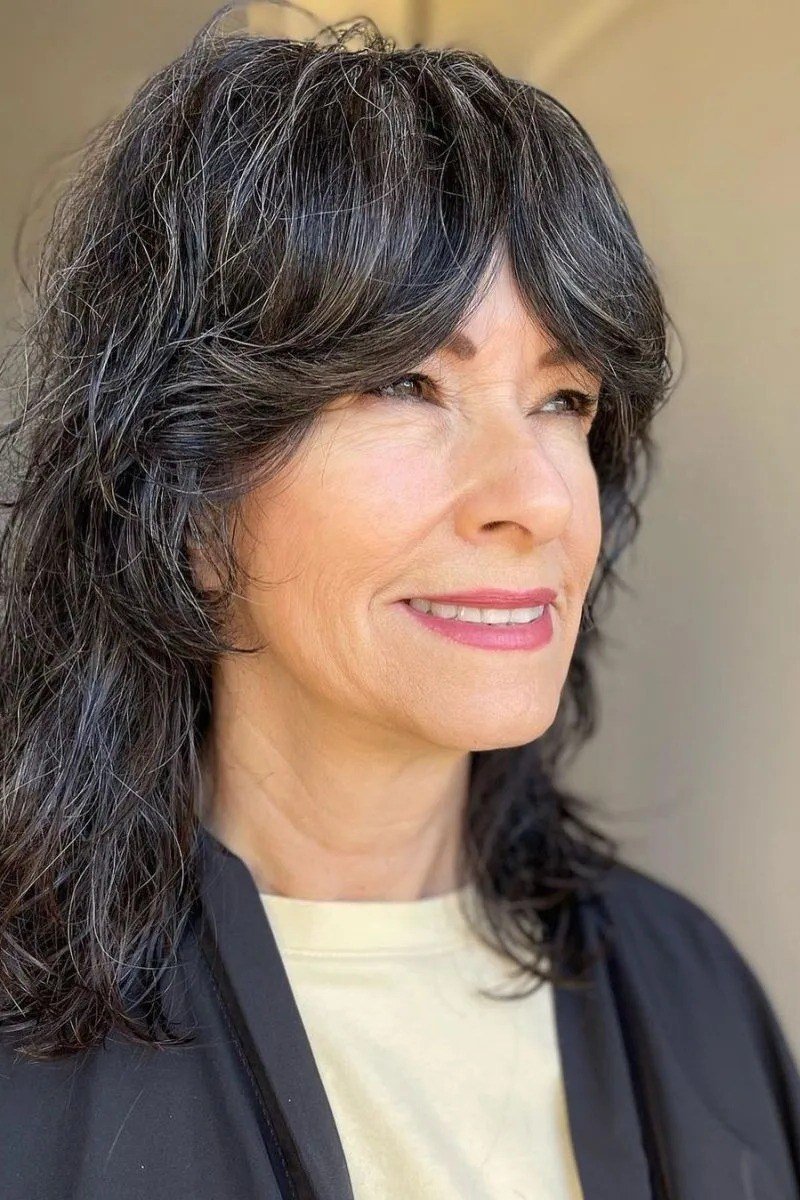 long salt-and-pepper wavy hair with bangs, three-quarter view, low-maintenance hairstyle for women over 40