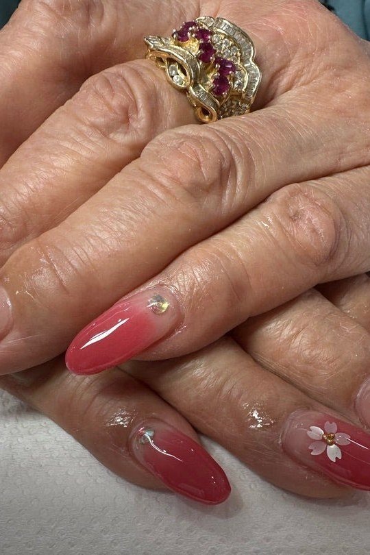 Red-to-pink ombre almond nails with tiny gold dots and a small floral accent, mature hands