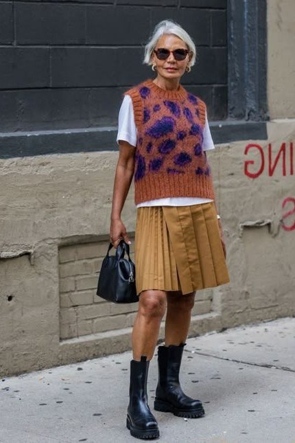 Woman over 40 wearing a purple knit vest over a white tee with a mustard pleated skirt