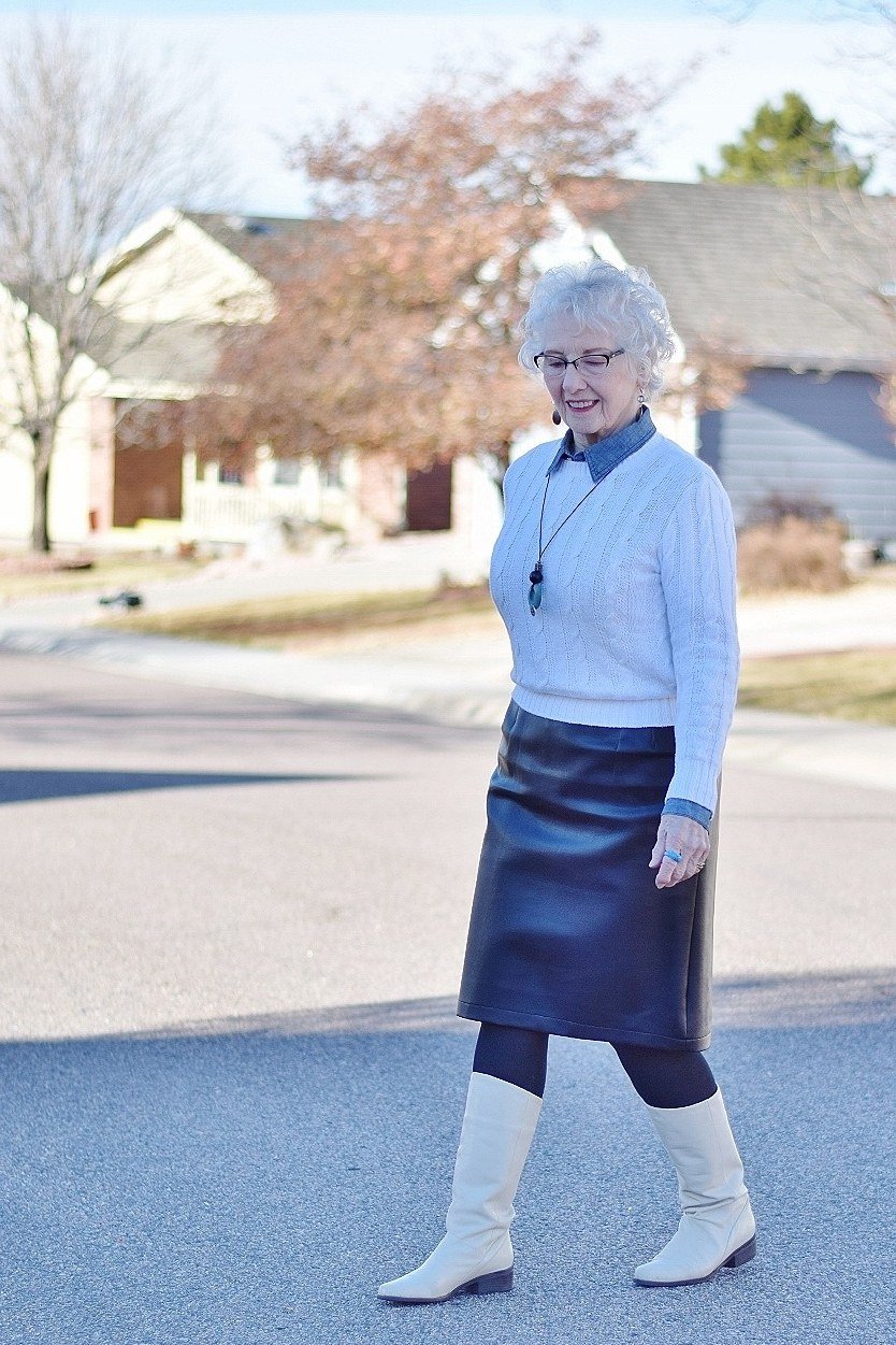 Woman over 40 wearing a light blue sweater with a navy leather midi skirt and white boots
