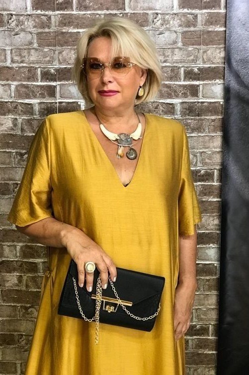 Effortlessly polished outfit for women over 50: A gold satin blouse with statement necklace, holding a black quilted clutch...