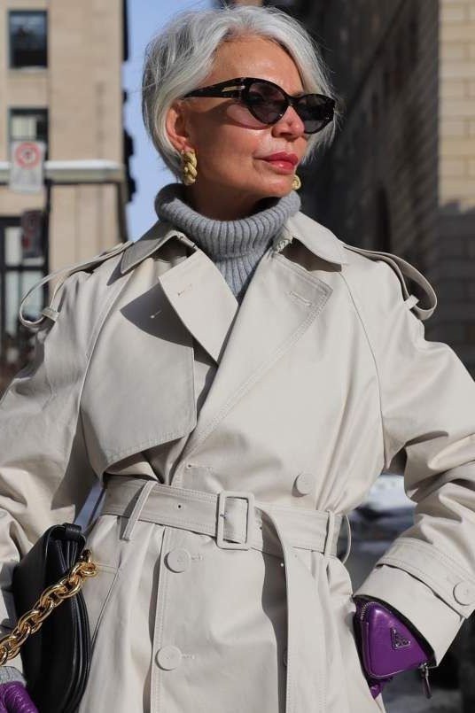 Effortlessly polished outfit for women over 50: A light stone trench coat over a gray turtleneck, carrying a black quilted...