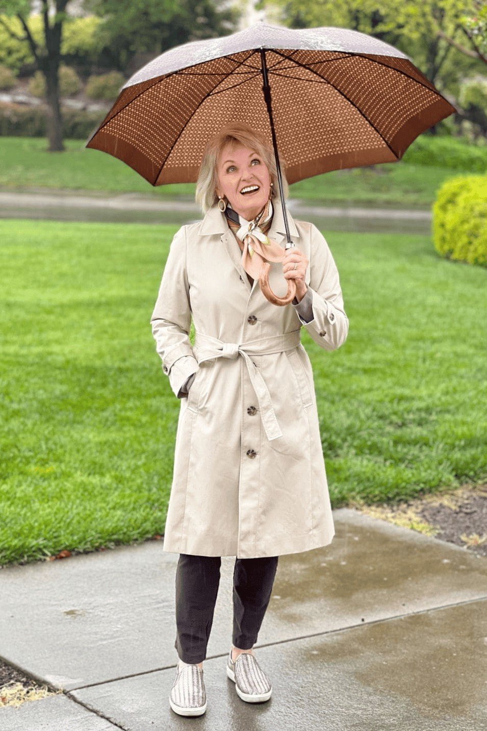 Effortlessly polished outfit for women over 50: Holding a tan umbrella, a light beige trench coat and light jeans, white...