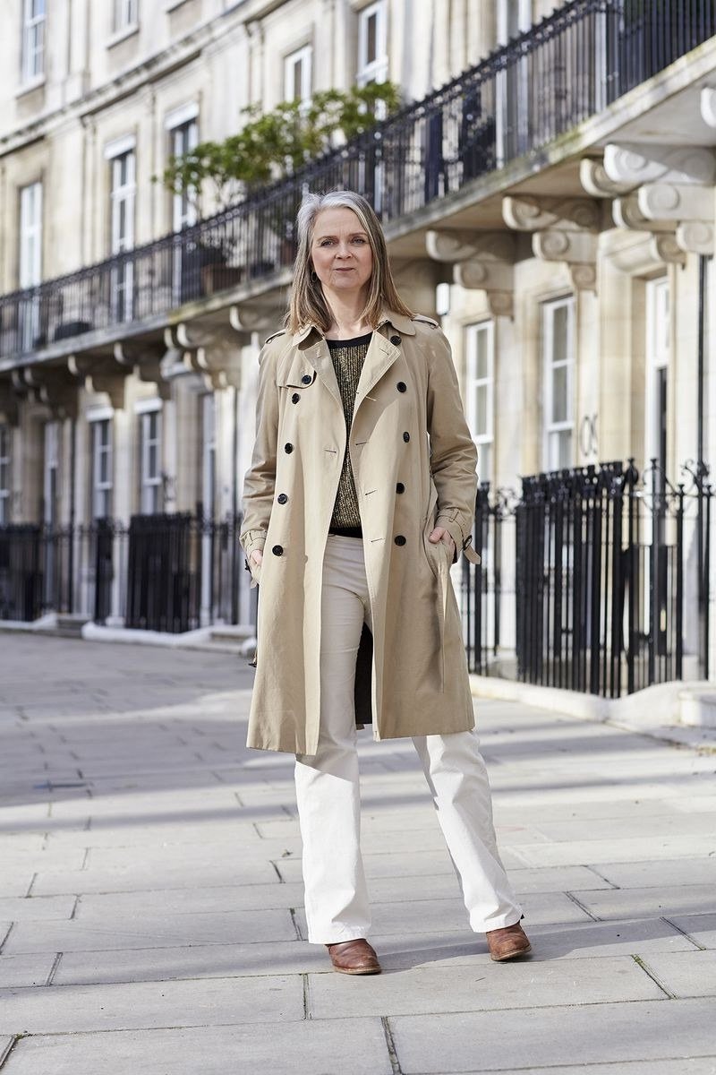 Woman over 70 in a classic beige trench coat over dark top and white trousers on a city street