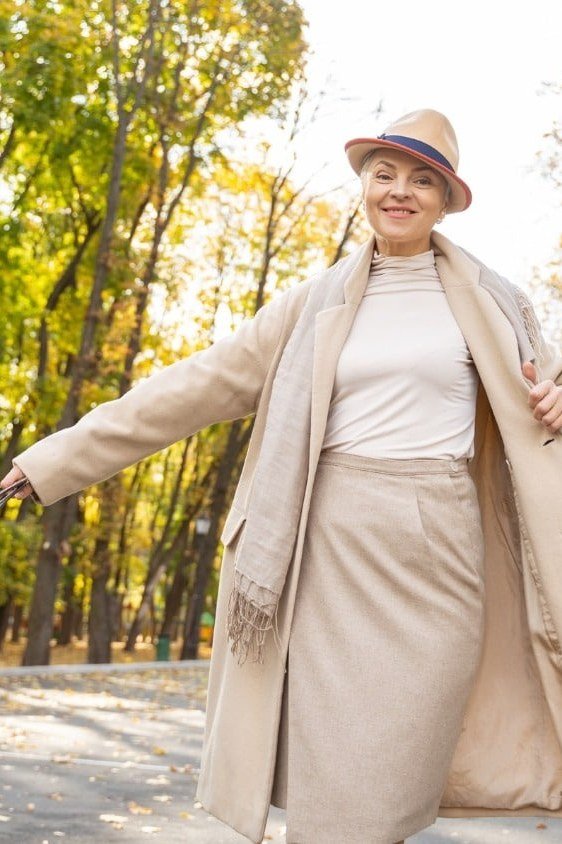 Woman over 70 in a beige trench coat, cream top and ivory skirt with a fedora in a fall park
