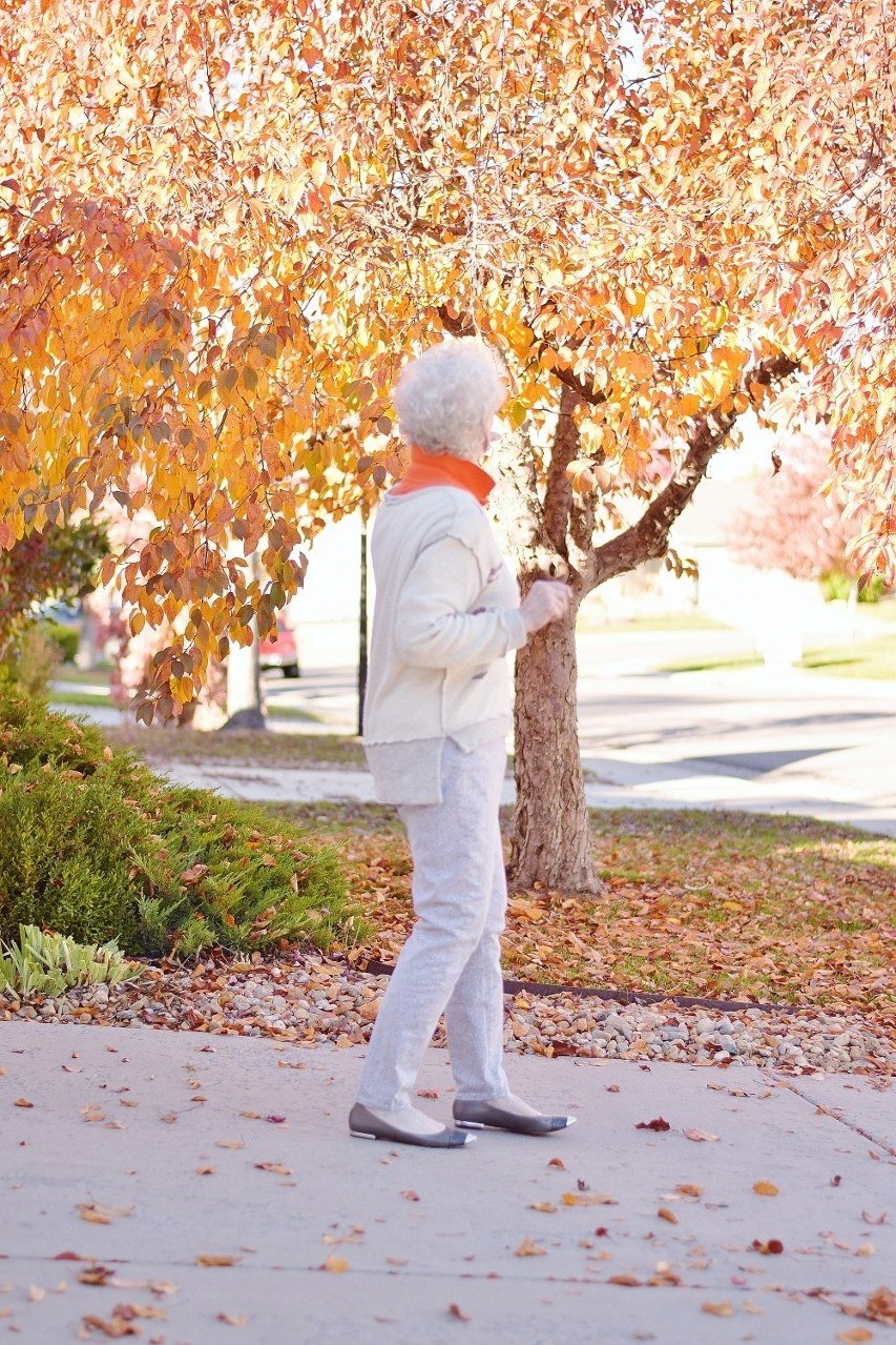 Older woman in an all-white outfit with a coral scarf strolling under fall trees