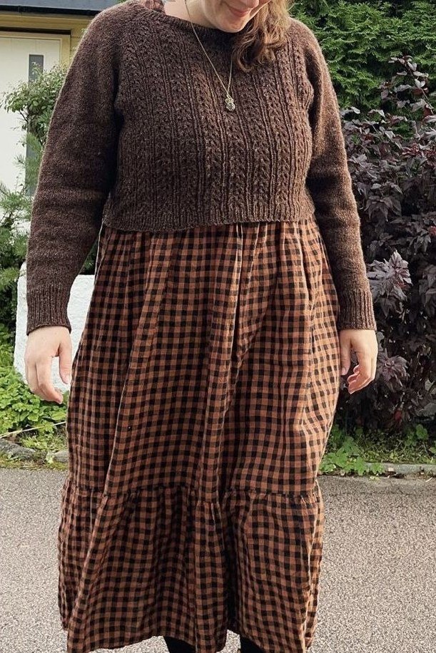 Woman over 70 wearing a brown cable knit sweater with a brown check midi skirt in a garden