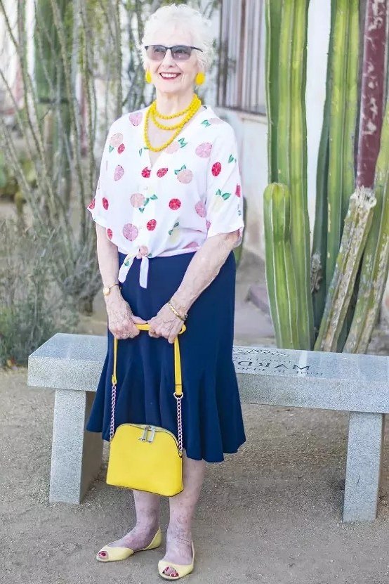 plus-size older woman wearing white cherry-print top with navy midi skirt and yellow handbag in garden