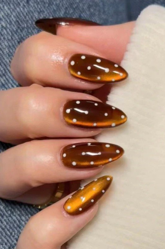Amber cat-eye almond nails with white polka dots beside a denim shirt cuff