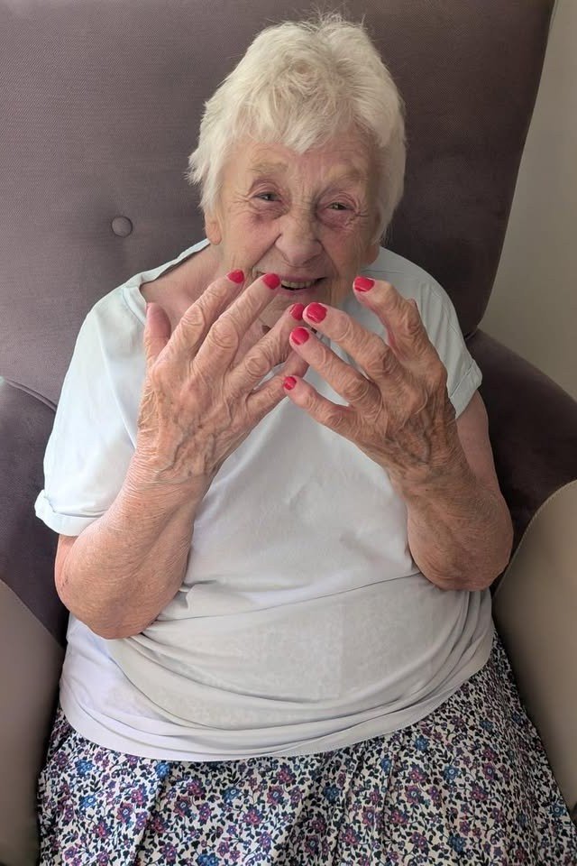 Elderly woman seated in an armchair showing bright red nails