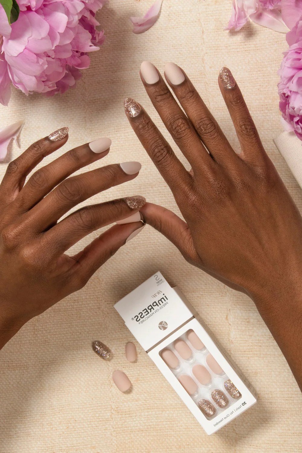 Mature dark-skinned hands with nude nails beside an imPRESS press-on box and flowers