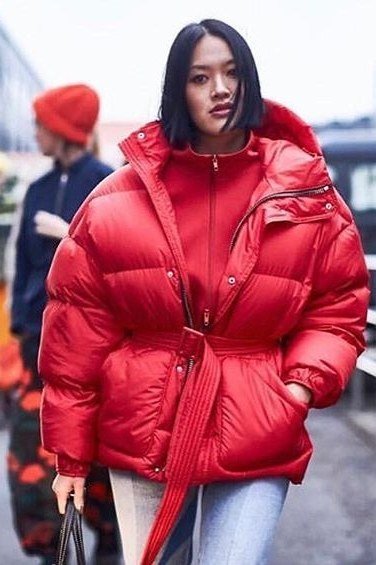 Woman over 60 wearing a red belted glossy puffer coat over a black top with blue jeans, street style