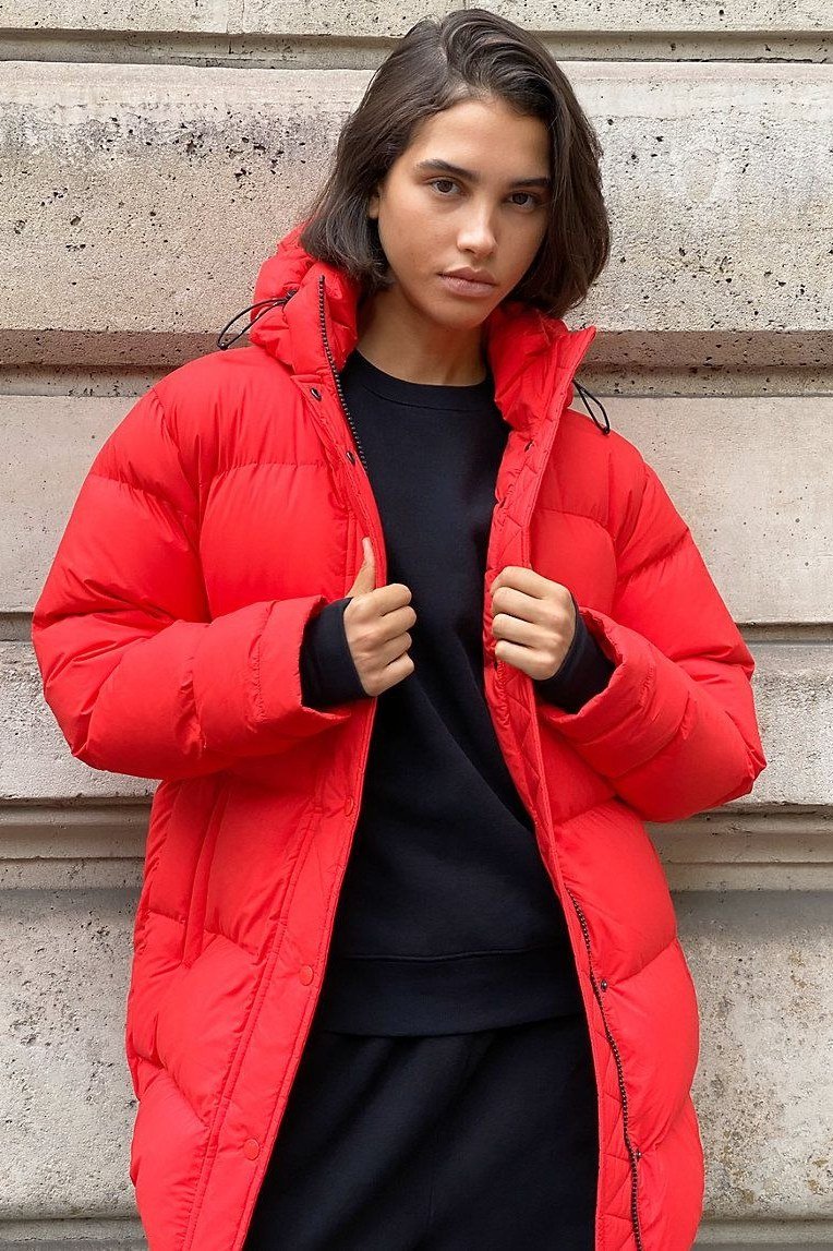 Woman over 60 wearing a red puffer coat over a black top, standing against a stone wall