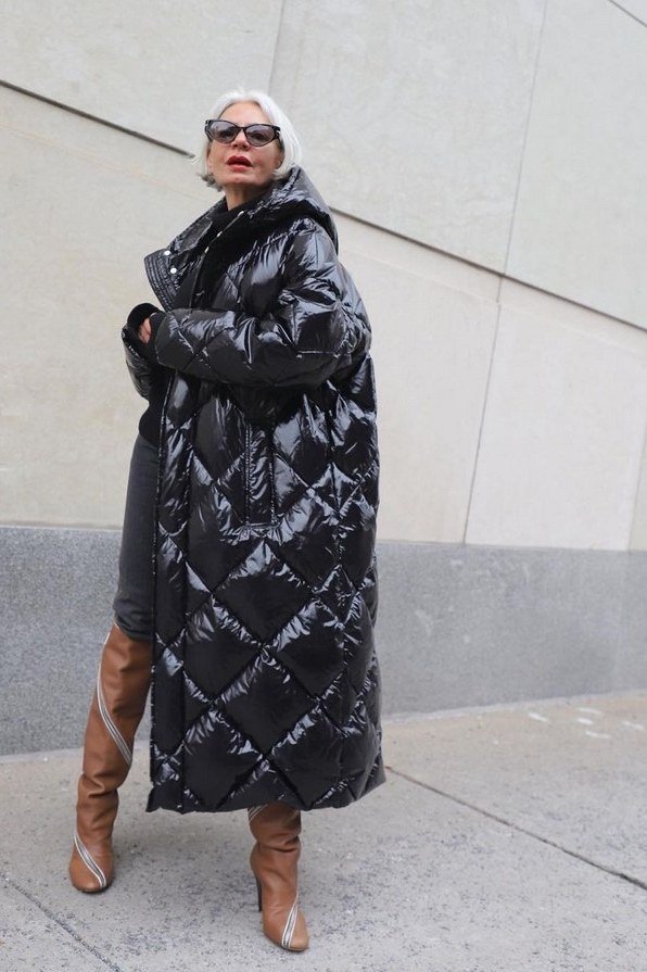 Woman over 60 wearing a long black diamond-quilted puffer coat with tan knee boots