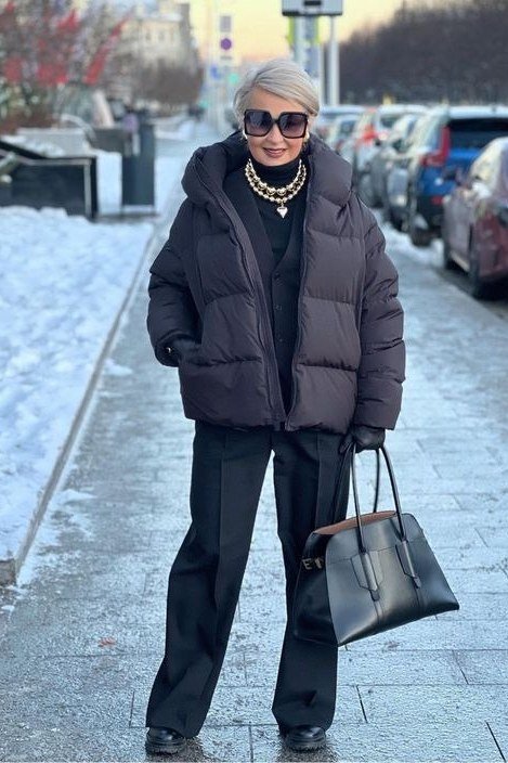 Woman over 60 wearing a black puffer coat over black wide-leg trousers on a snowy street