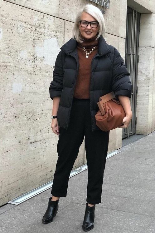 Woman over 60 wearing a black puffer coat over a brown turtleneck with black cropped pants and tan bag