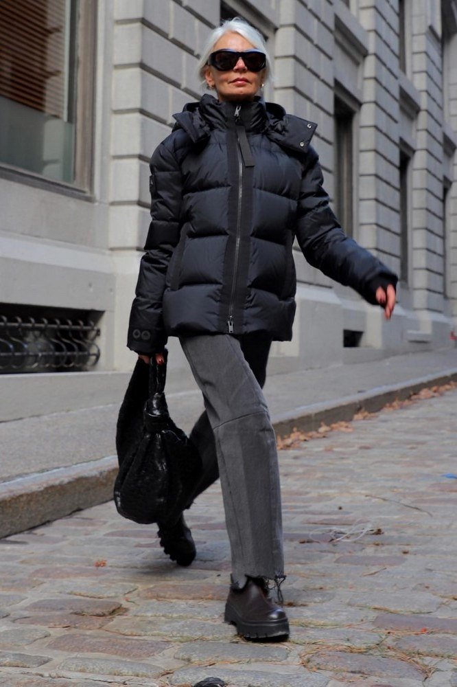 Woman over 60 wearing a black puffer coat with gray trousers and black bag on a city street