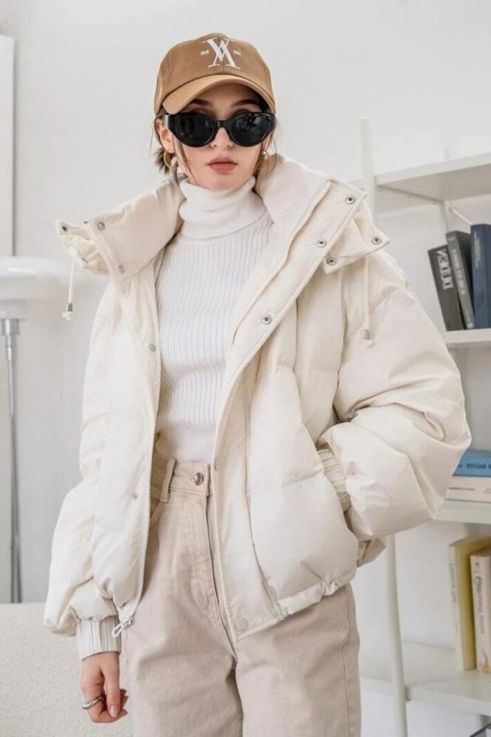 Woman over 60 wearing a cream puffer coat with a baseball cap over a white top and beige pants
