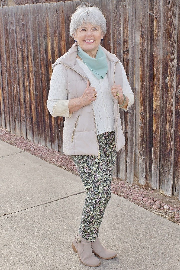 Woman over 60 wearing a cream puffer coat with patterned leggings and ankle boots by a wooden fence