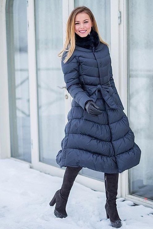 Woman over 60 wearing a long belted navy puffer coat with black boots on a snowy street