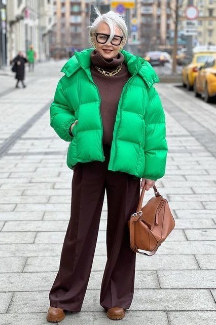Woman over 60 wearing a green puffer coat with brown trousers and tan bag on a city street