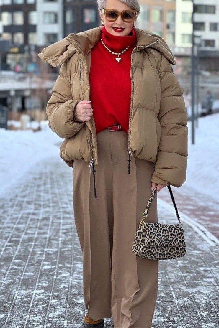 Woman over 60 wearing a camel puffer coat over a red turtleneck with tan wide-leg trousers and leopard bag in the snow