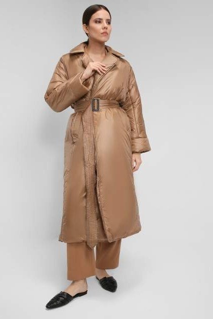 Woman over 60 wearing a camel belted long quilted coat over tan trousers