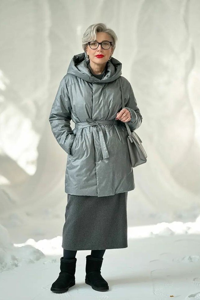 Woman over 60 wearing a gray belted puffer coat over a long gray skirt with black boots