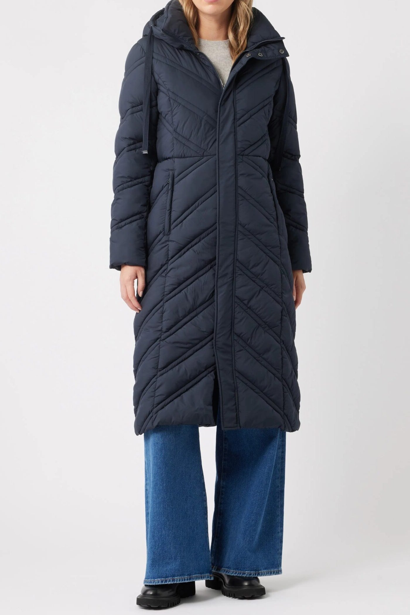 Woman over 60 wearing a navy chevron-quilted long puffer coat over wide blue jeans